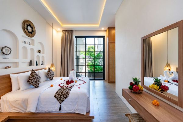 Romantic tropical bedroom with king bed decorated in rose petals and swan towels, wooden accents, fruit display, and garden-view sliding doors.